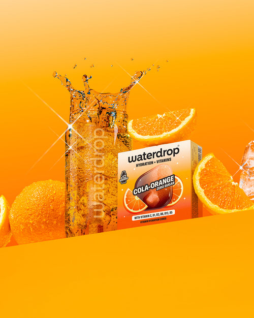 COLA ORANGE Vitaminwürfel mit sprudelndem Wasserglas und frischen Orangen.