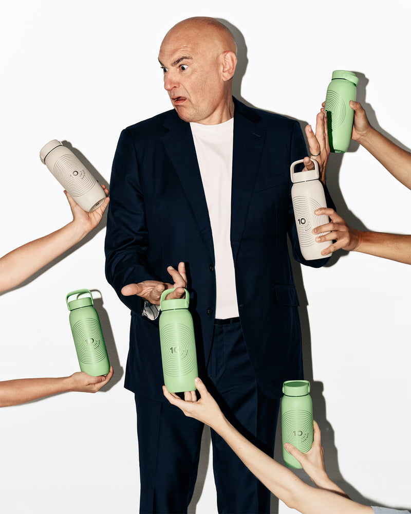 Ein Mann in dunklem Anzug steht, während mehrere Hände grüne und weiße Trinkflaschen um ihn herum reichen.
