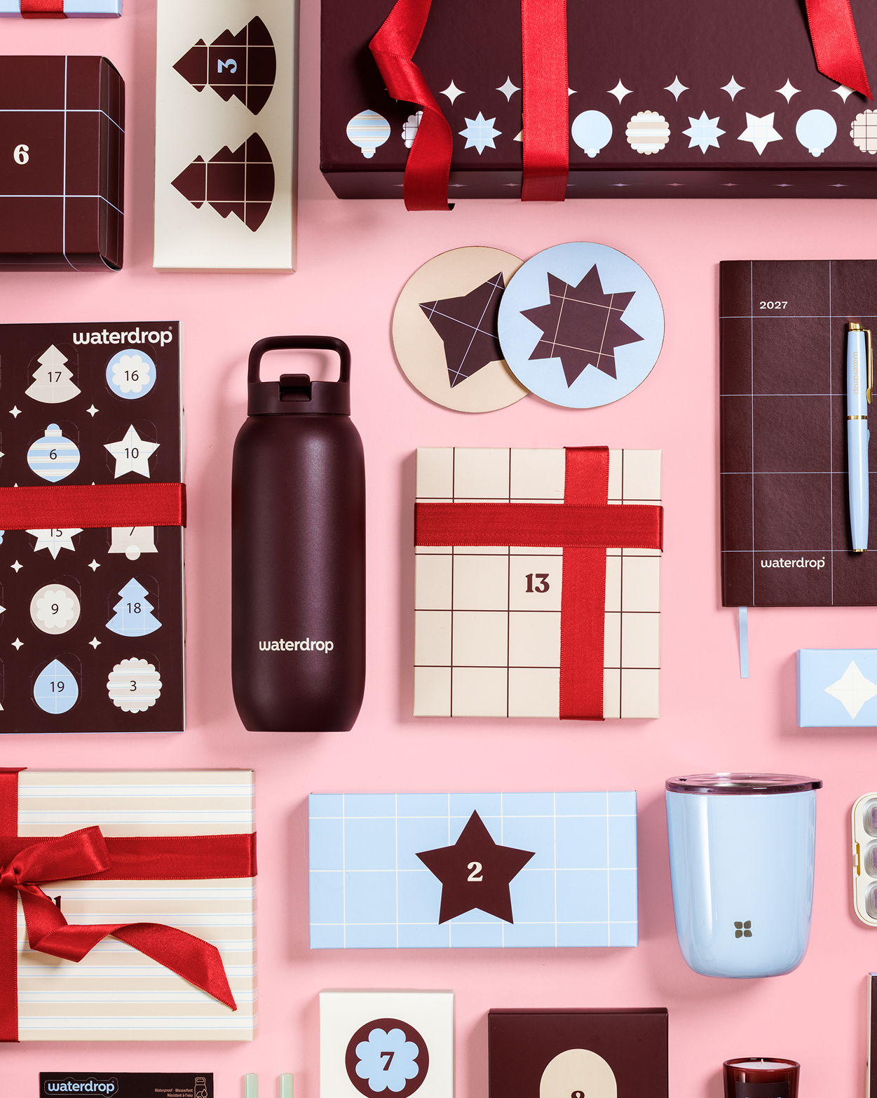 Verschiedene Geschenkboxen, ein Adventskalender, eine Wasserflasche, eine Tasse und weitere verpackte Geschenke auf rosa Hintergrund.