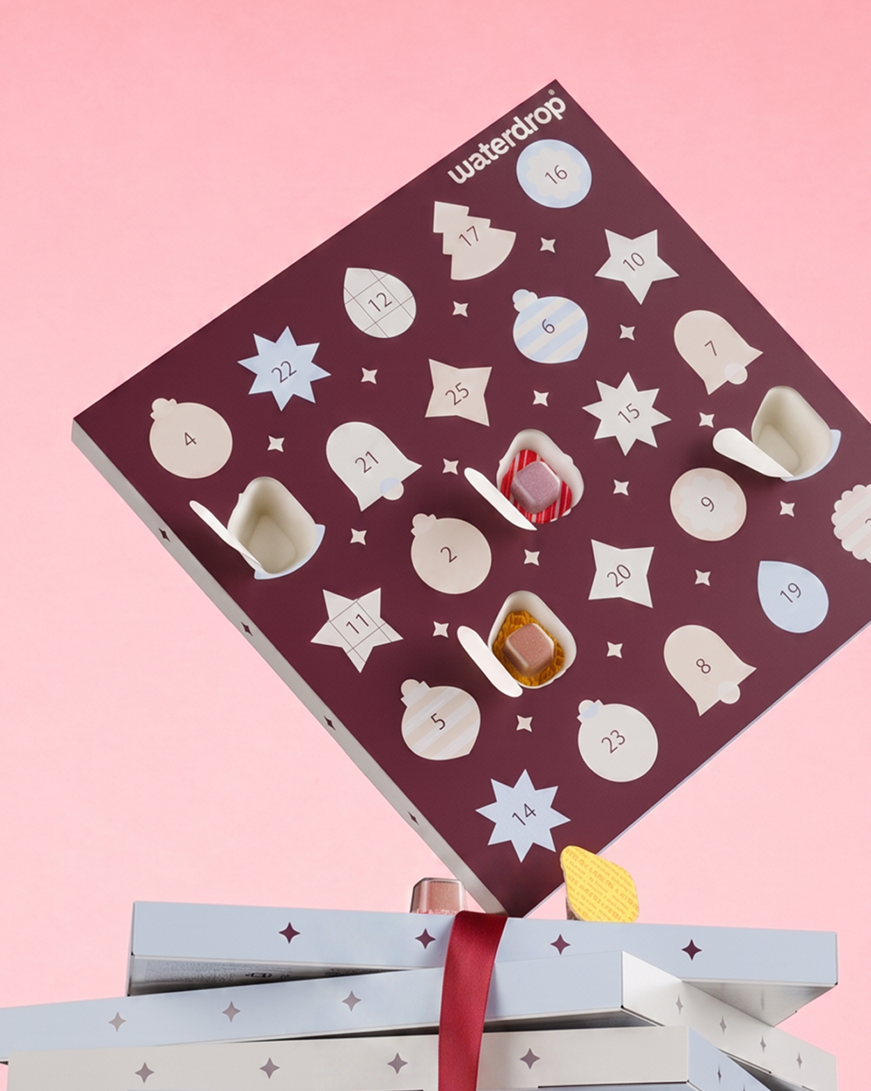 Lila Adventskalender mit nummerierten sternförmigen Türen vor rosa Hintergrund, über zwei verpackten Geschenken.