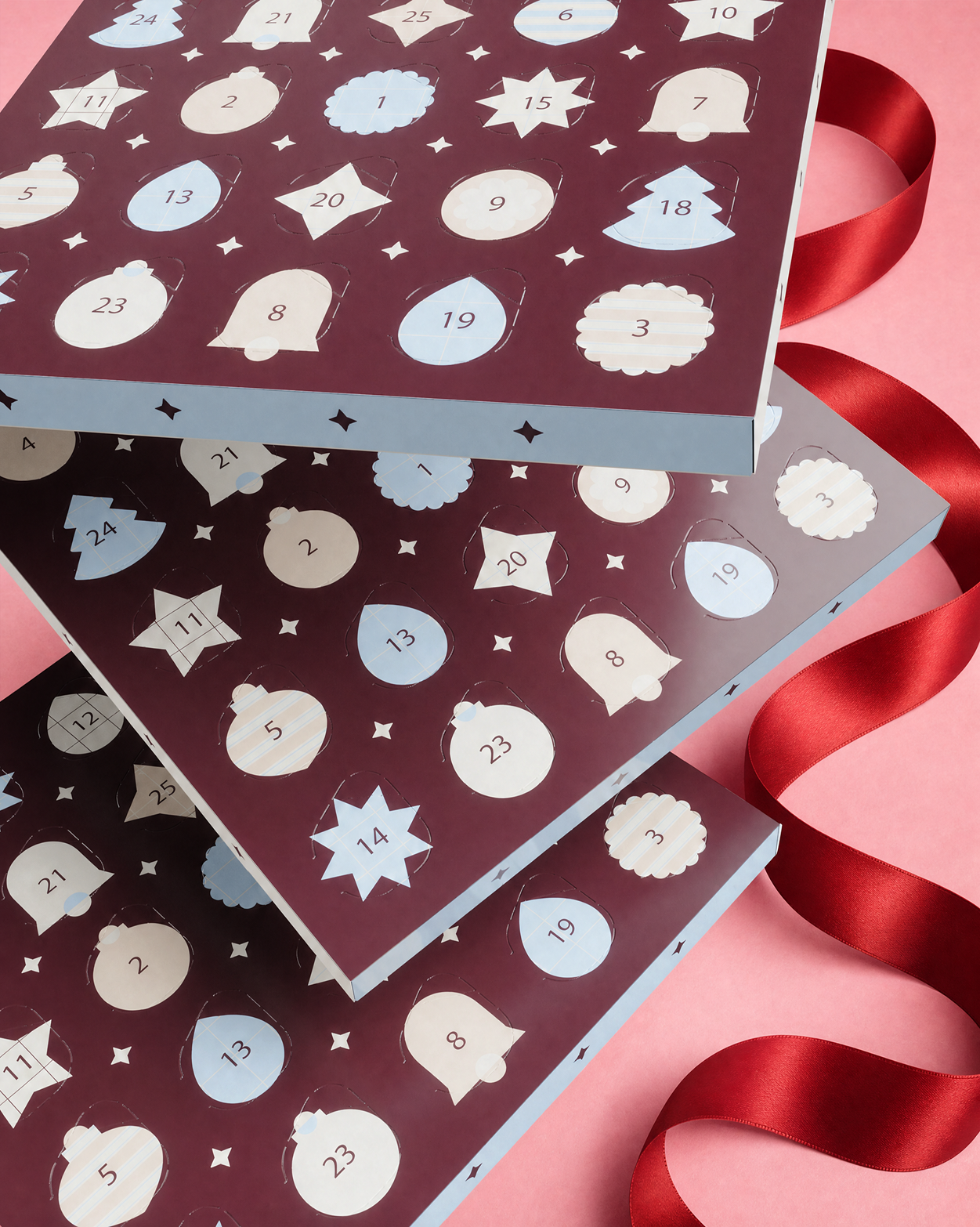Drei stapelbare Adventskalenderboxen mit nummerierten Türen und dekorativen Formen, mit roter Schleife auf pinkem Hintergrund.