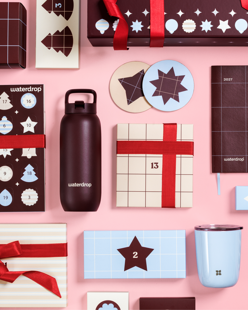 Verschiedene Adventskalender und Geschenkboxen mit roten Schleifen vor rosa Hintergrund, neben einer schwarzen Wasserflasche und einer hellblauen Tasse.