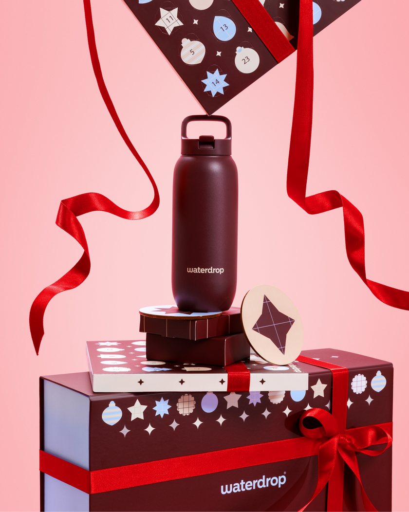 Schwarze Wasserflasche mit Deckel auf gestapelten Geschenkpaketen mit roten Bändern vor rosa Hintergrund.