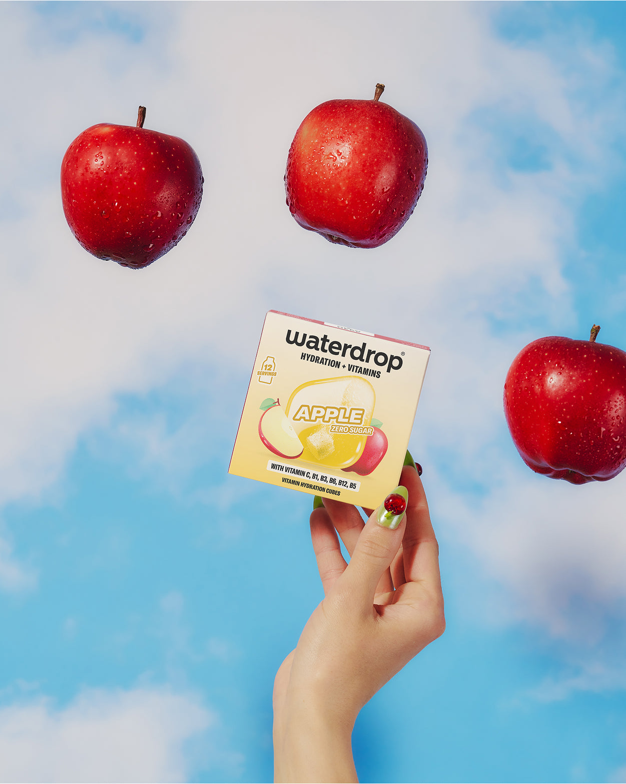 Eine Hand hält eine kleine Box mit einer Apfel-Illustration vor blauer Himmel, drei rote Äpfel schweben.