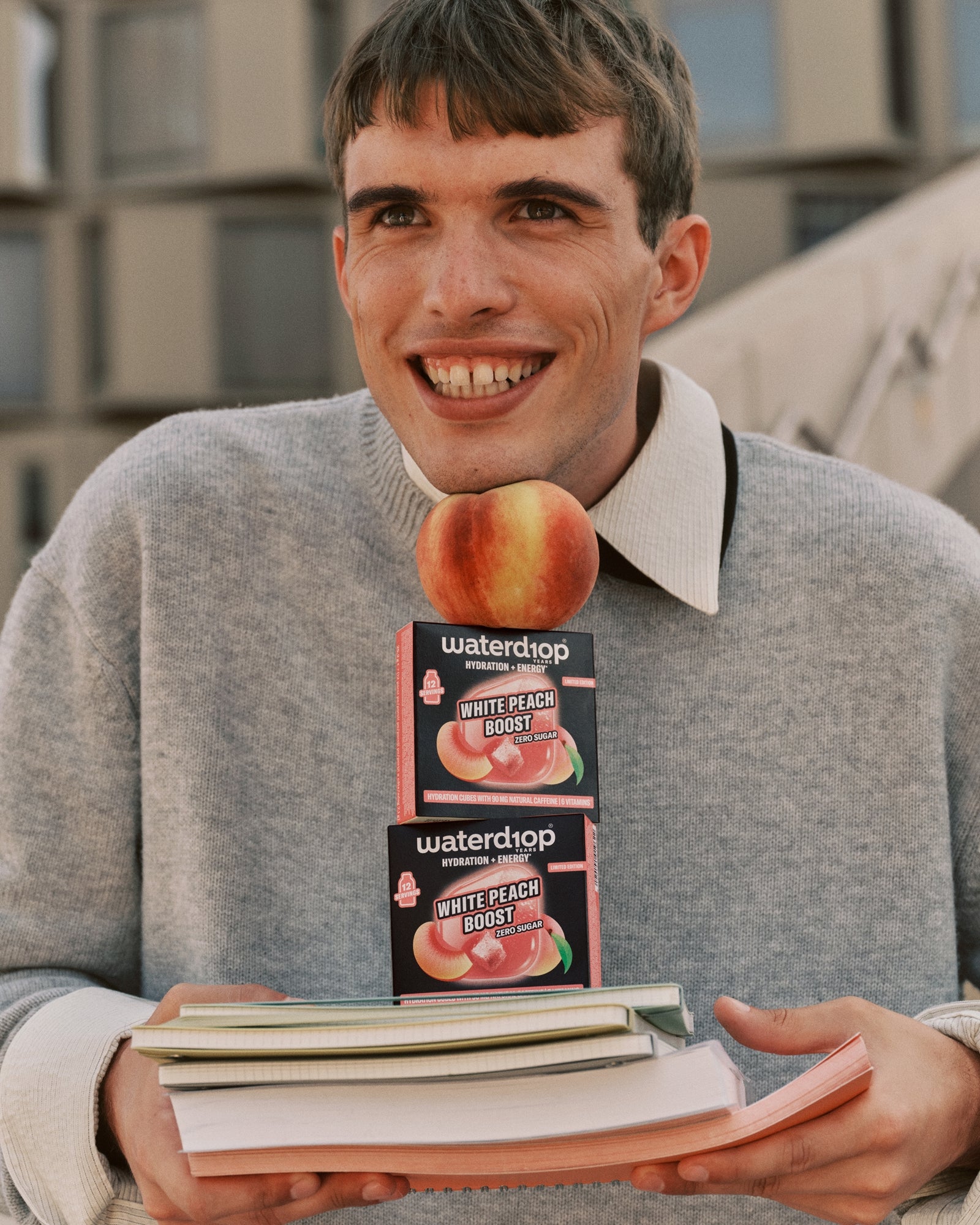 Junger Mann in grauem Pullover hält zwei Pfirsich-Geschmacksboxen auf einem Stapel Bücher.