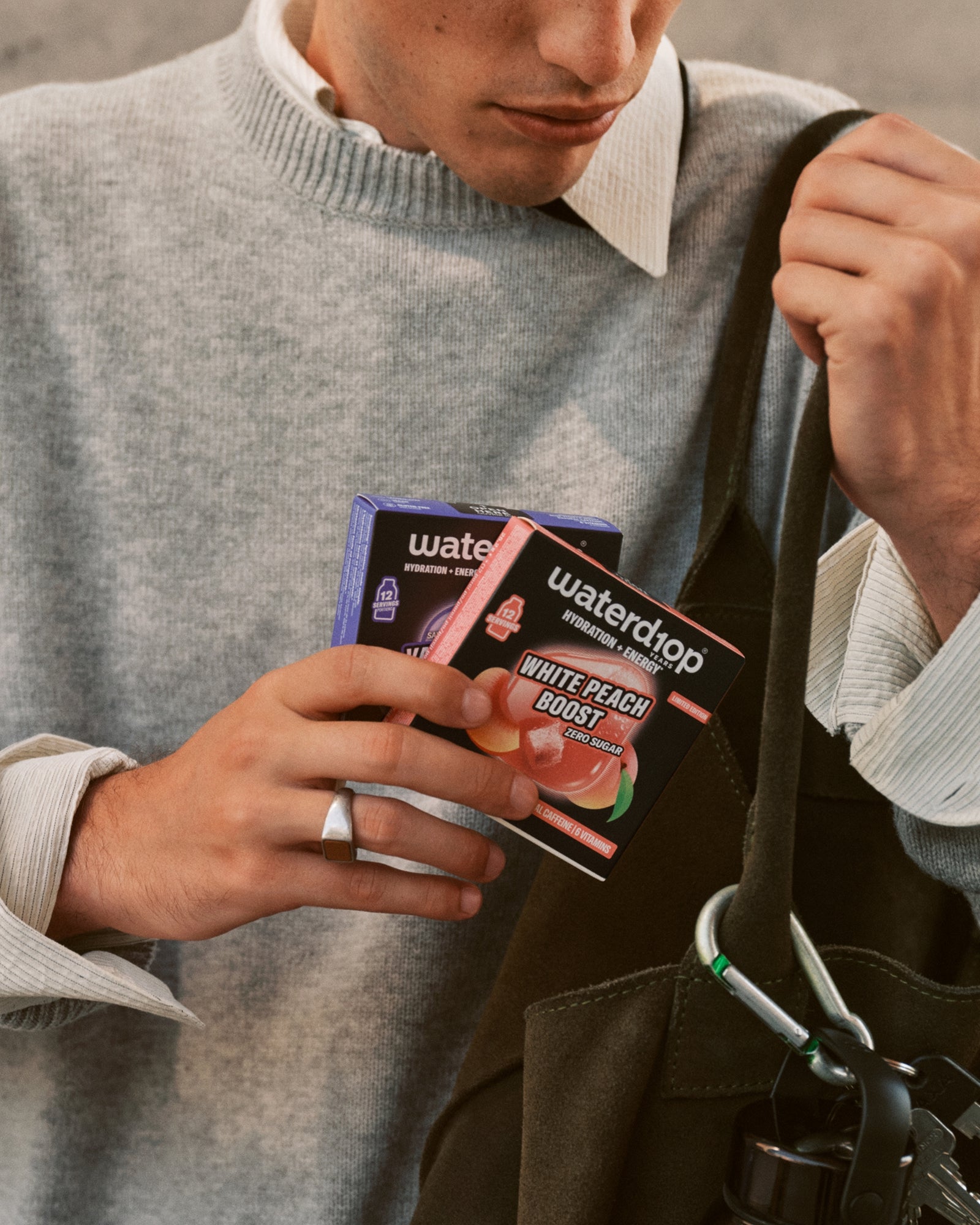 Person in grauem Pullover hält kleine Produktboxen neben einer schwarzen Tragetasche.