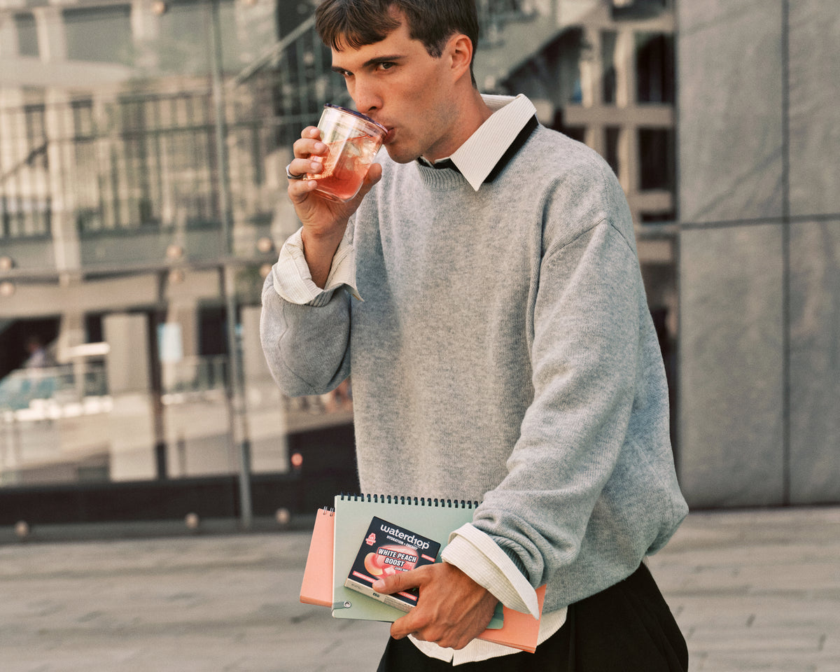Mann in grauem Pullover trinkt aus einem Glas und hält draußen in einer städtischen Umgebung ein Notizbuch.