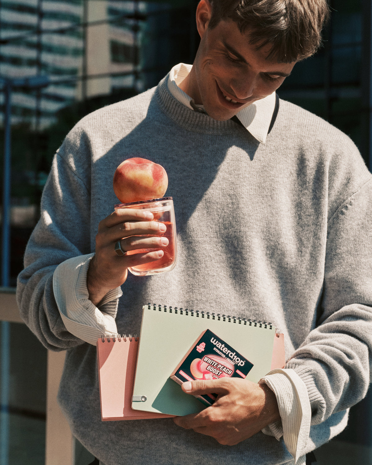 Mann in grauem Pullover steht draußen und hält ein rosafarbenes Getränk mit Apfel am Rand sowie ein Notizbuch in der anderen Hand.