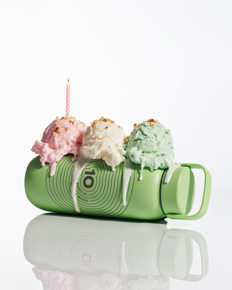 Grüner Becher liegt seitlich, drei Kugeln Eis oben und eine brennende Kerze, schmilzender Zuckerguss tropft herab.