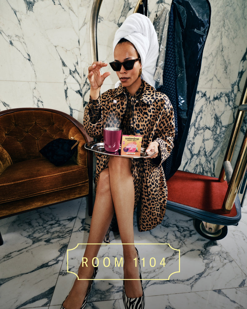 Frau in Leopard-Print-Kimono mit Handtuch auf dem Kopf sitzt auf Marmorboden und hält ein Tablett mit Kosmetikprodukten.
