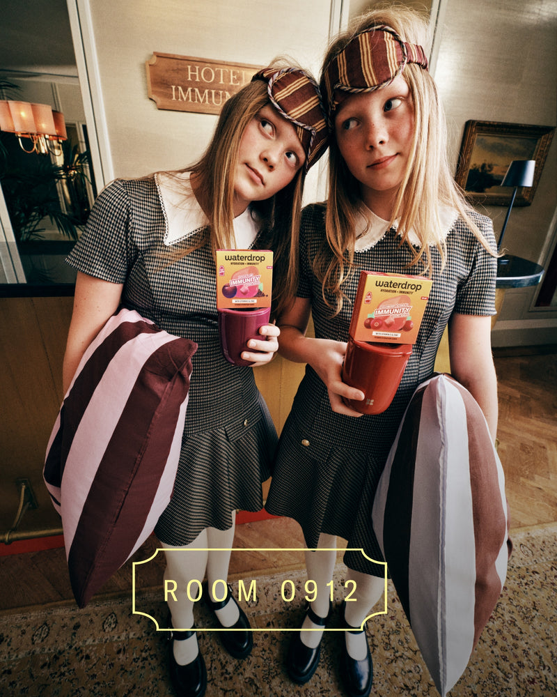 Zwei Mädchen in passenden Outfits mit karierter Stirnlampe halten bunte Getränke in der Hotellobby, stehen nebeneinander mit gestreiften Taschen.