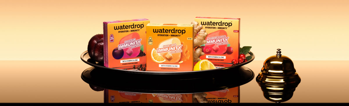 Tablett mit drei Waterdrop-Verpackungen auf einer reflektierenden Oberfläche und einer kleinen dekorativen Flasche rechts.