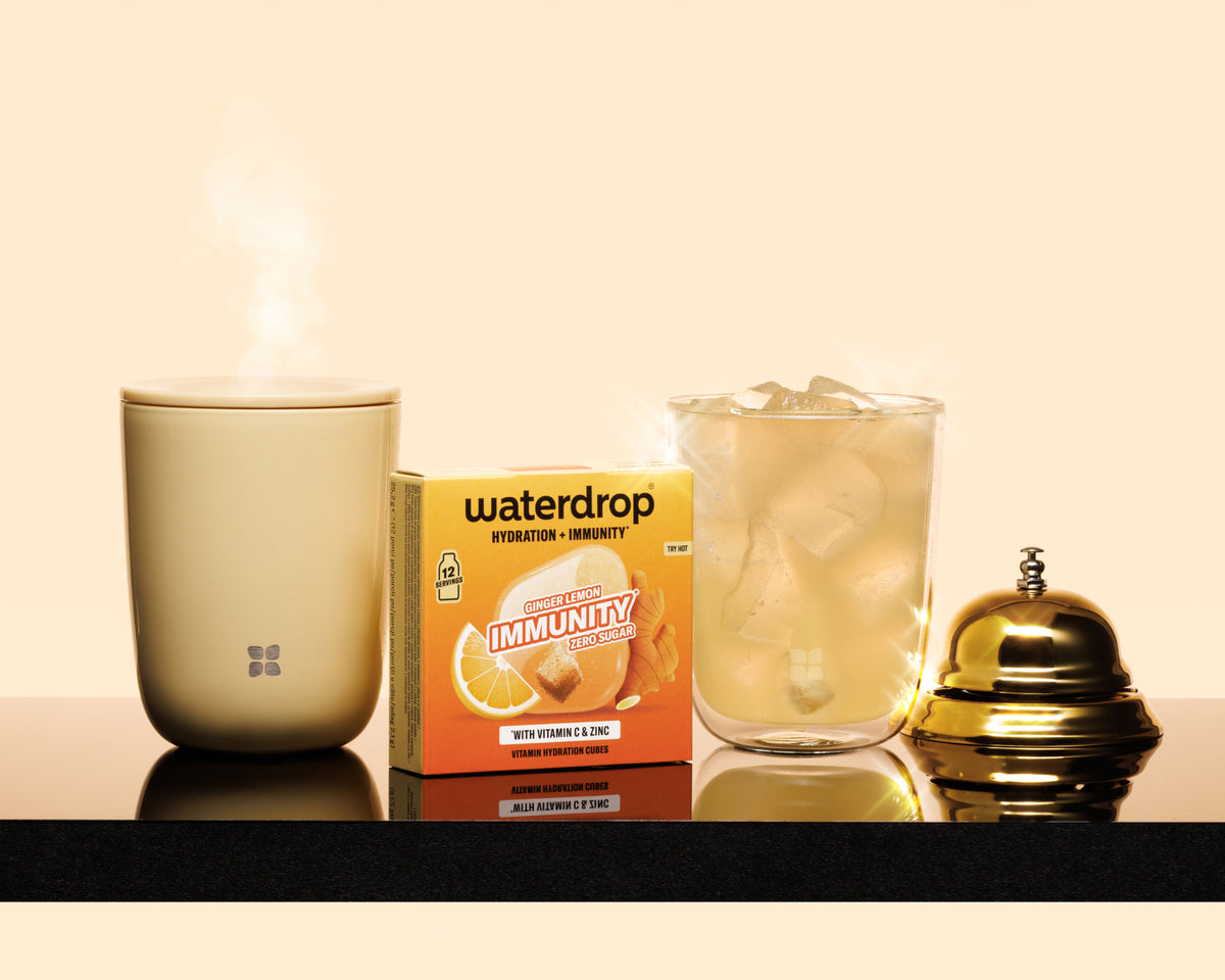 Dampfende beigefarbene Tasse, eine Immunitätsbox, ein Glas mit Eis und eine goldene Glocke auf einer schwarzen Arbeitsfläche.