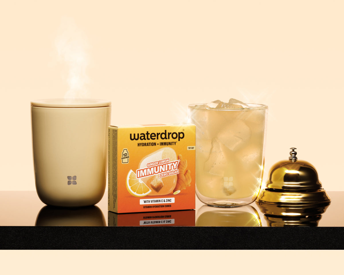 Beige dampfender Becher, eine Verpackungskiste, ein Glas mit Eiswürfeln und eine Messingglocke auf einem dunklen Regal.
