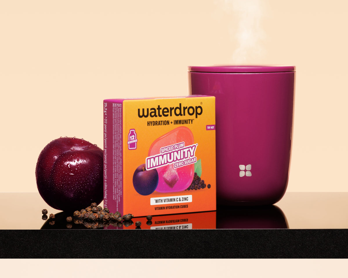 Violette Tasse mit Dampf neben einer orangefarbenen Waterdrop-Box, eine Pflaume und verstreute Samen auf einer spiegelnden Oberfläche.