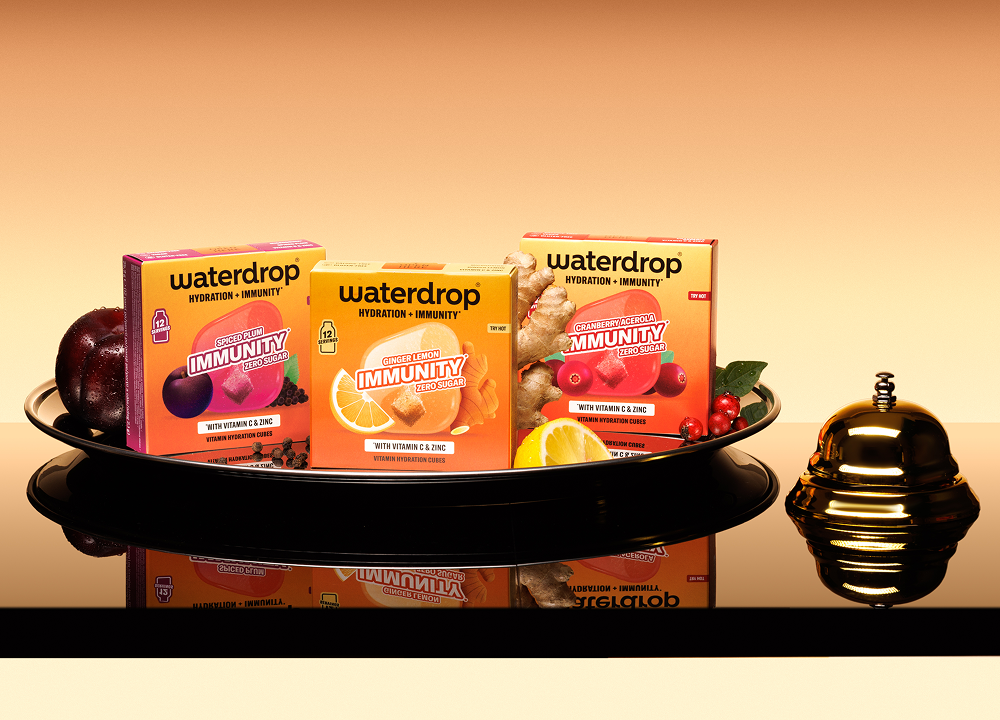 Verschiedene Getränkepackungen auf einer schwarzen Platte mit reflektierender Oberfläche und einer kleinen goldenen Glocke rechts.