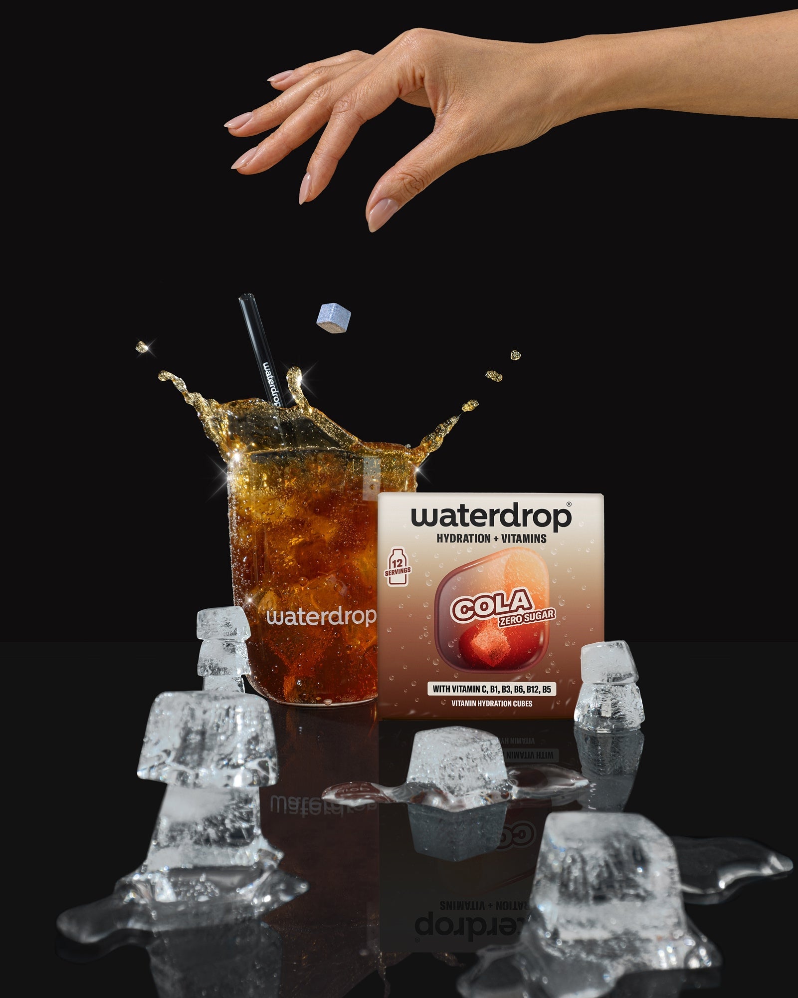 Eine Hand reicht über ein Glas Cola mit Eiswürfeln, dahinter steht eine Getränke-Verpackung.