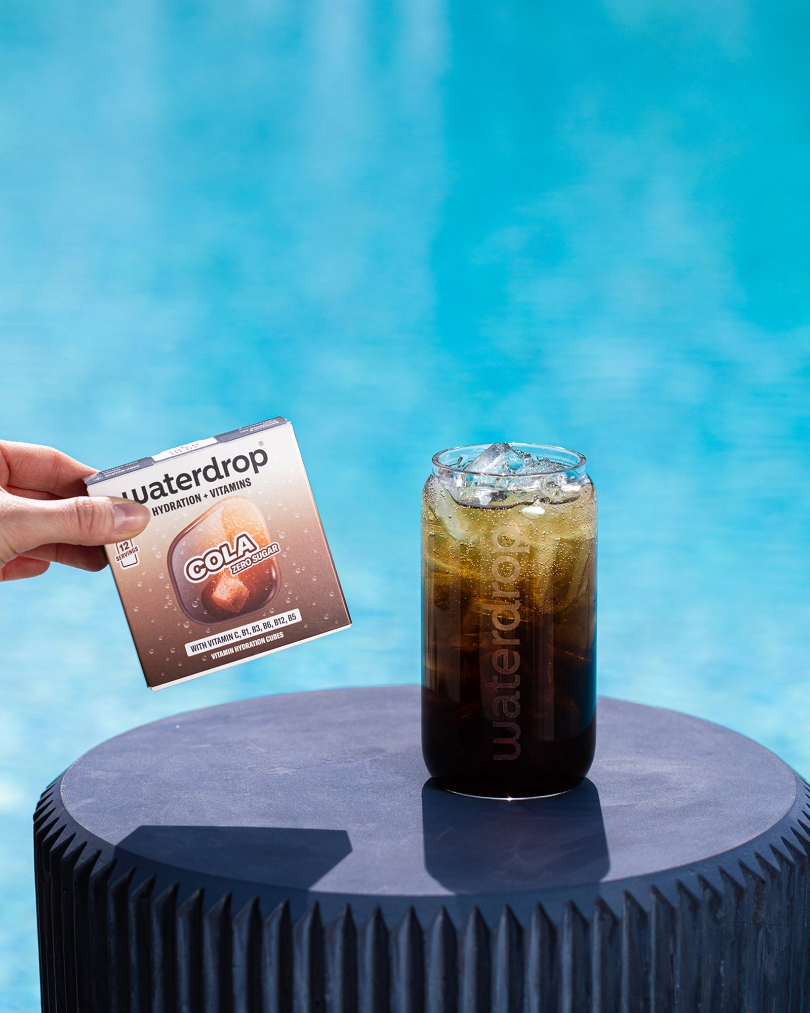 Hand hält ein kleines Paket neben einem Glas mit eiskalter Cola auf einem runden blauen Podest am Pool.